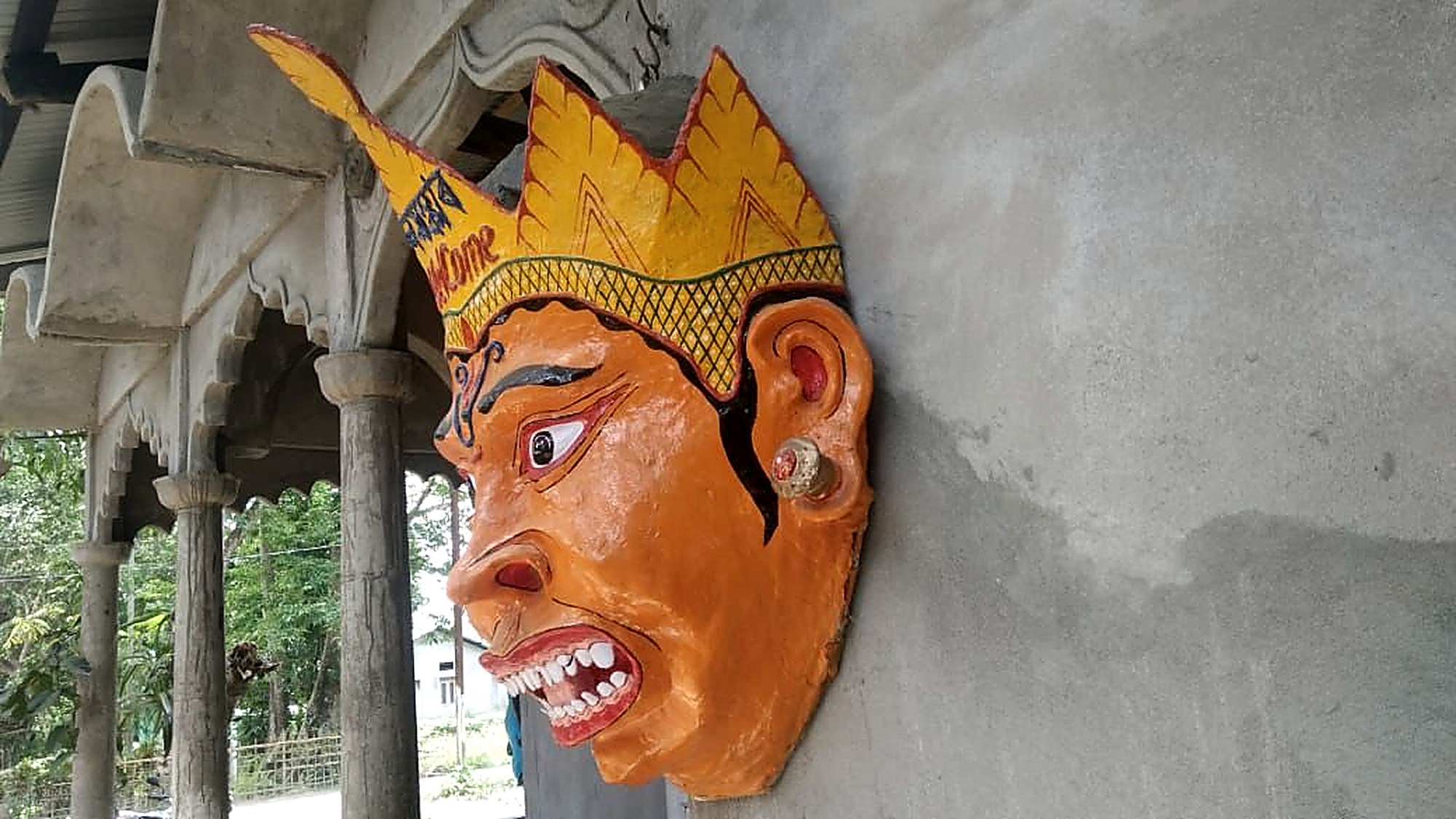 Large ochre mask mounted on a namghar wall, greeting visitors in a village in Assam
