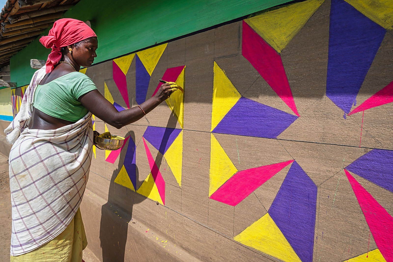 A woman painting a large geometric mural in pink, purple and yellow on a village wall