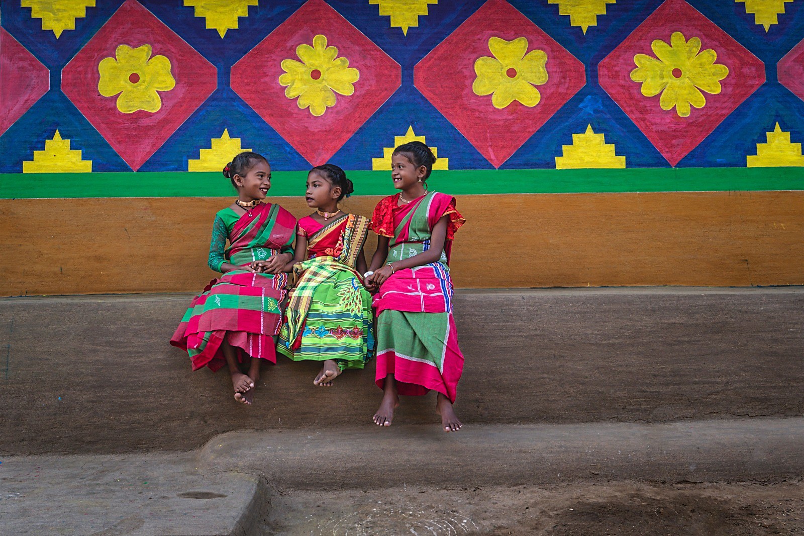 Three girls in bright traditional dress seated before a painted village wall
