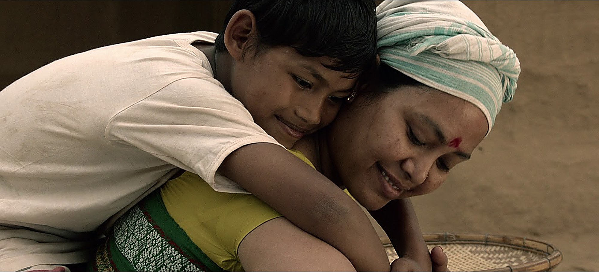 Film still: a boy resting on his mother's shoulder in a village courtyard