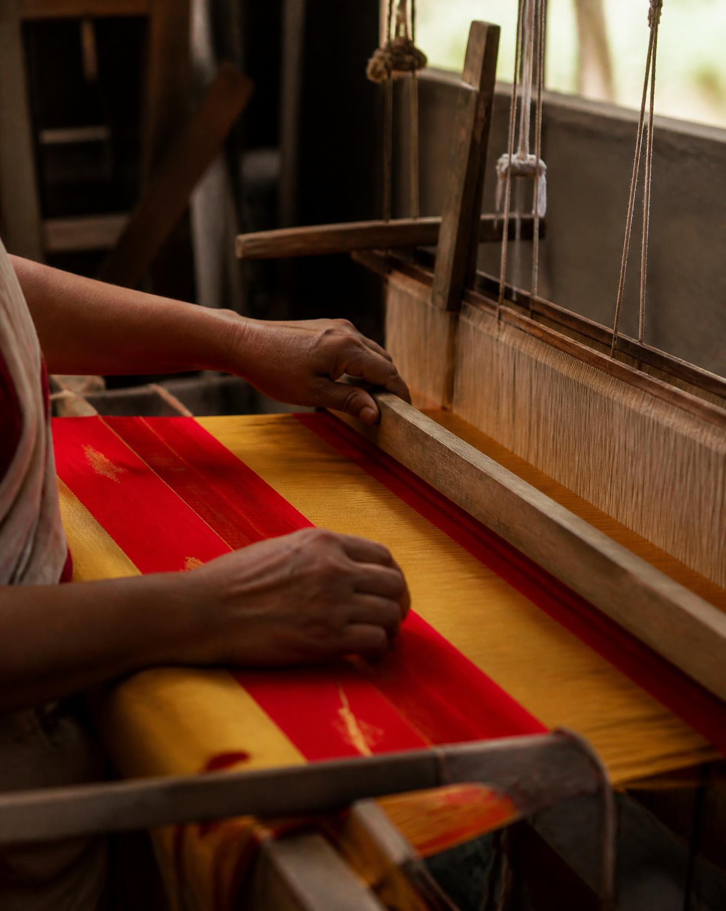 Hands weaving red and gold silk on a bamboo loom